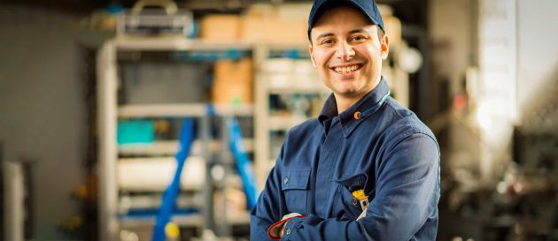 Lächelnder Industriemechaniker in blauer Arbeitskleidung steht mit verschränkten Armen in einer Werkstatt.