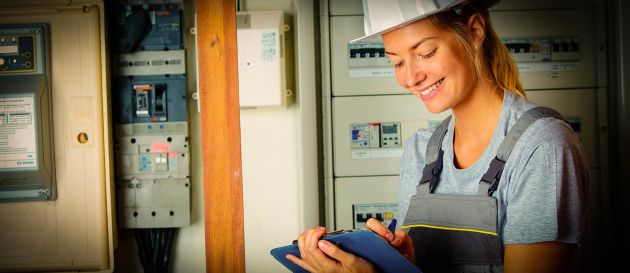Junge Elektrikerin mit Schutzhelm und grauer Arbeitskleidung steht lächelnd vor einem Schaltschrank und macht Notizen auf einem Klemmbrett.
