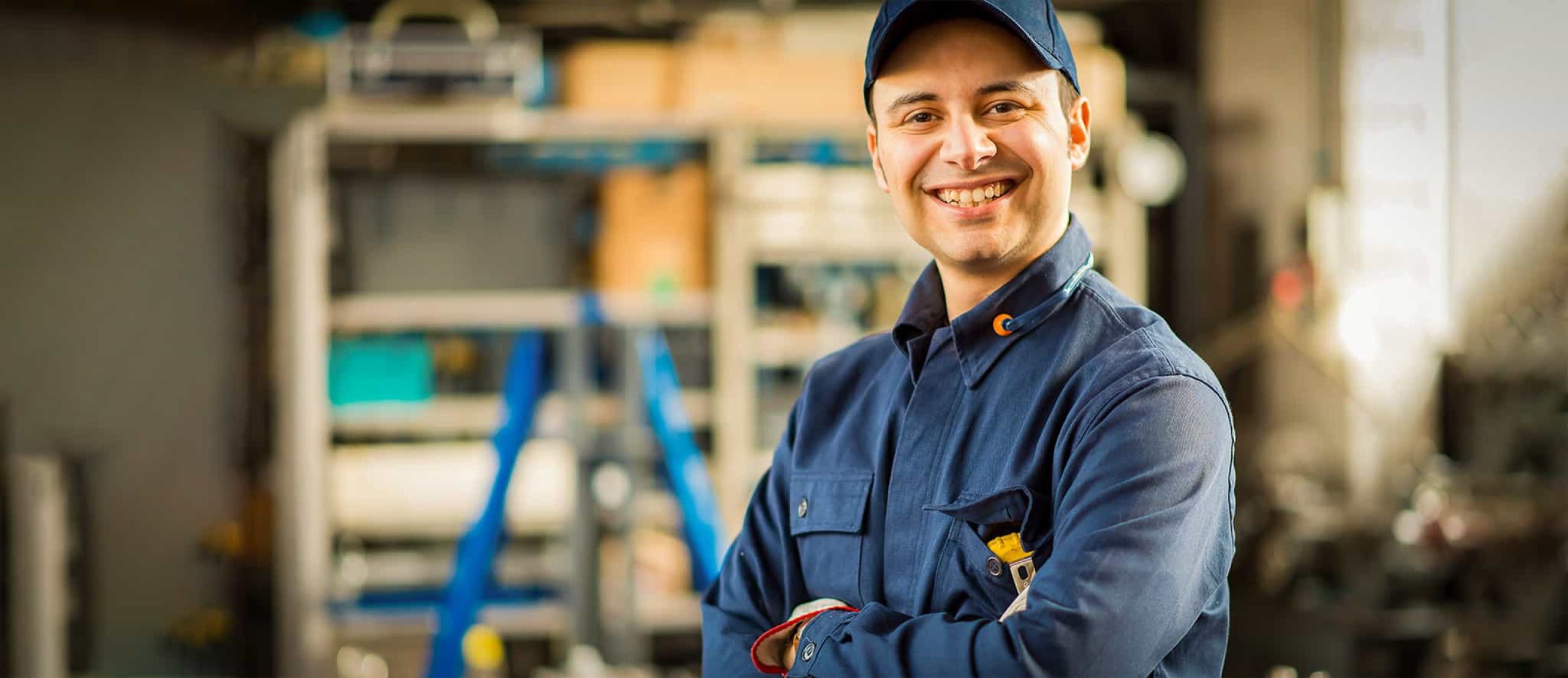 Lächelnder Industriemechaniker in blauer Arbeitskleidung steht mit verschränkten Armen in einer Werkstatt.