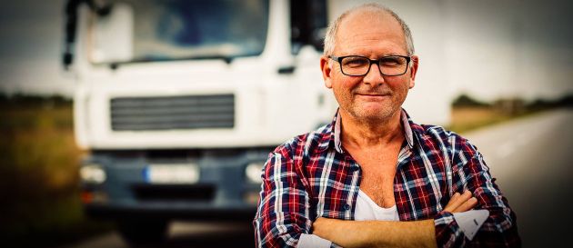 Ein älterer Mann mit Brille und kariertem Hemd steht mit verschränkten Armen vor einem weißen Lastwagen auf einer Landstraße. Er lächelt leicht in die Kamera.