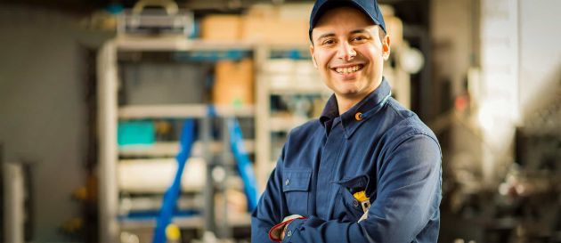 Lächelnder Industriemechaniker in blauer Arbeitskleidung steht mit verschränkten Armen in einer Werkstatt.